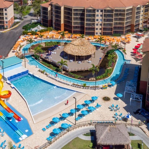 An aerial view showcases a resort pool area with water slides, lounge chairs, a tiki hut, and surrounding buildings.