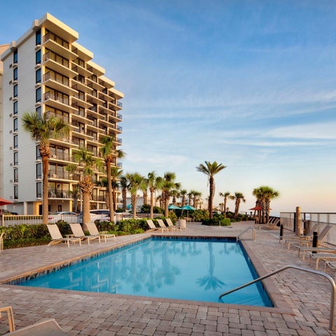 A rectangular pool sits adjacent to a multi-story hotel building with palm trees and a beach visible in the background.
