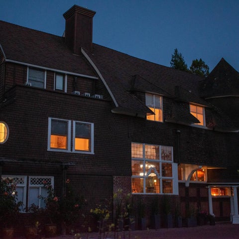A large, dark-colored building with multiple windows and a prominent turret is illuminated at dusk.
