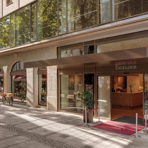 The entrance of Excelsior Hotel shows a modern facade with a covered entryway, reception desk, and potted plants.