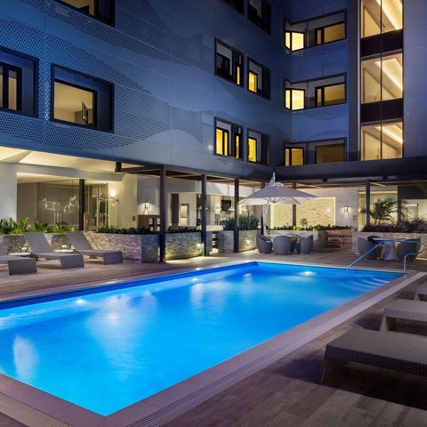A modern hotel features a rectangular swimming pool with lounge chairs and a patio area with greenery and outdoor lighting.