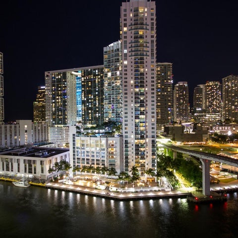 A nighttime view of a modern cityscape featuring illuminated skyscrapers, waterfront buildings, and a bridge.