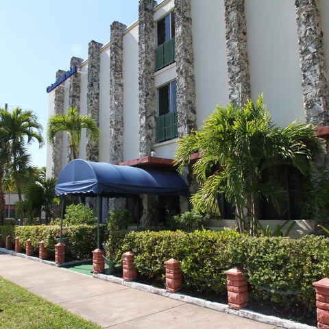 A building facade with a covered entrance, lush landscaping, and palm trees in a sunny outdoor setting.