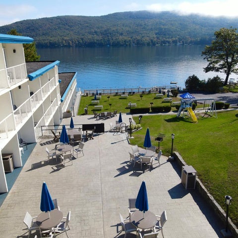 A hotel with a patio area featuring outdoor seating, umbrellas, and a playground overlooking a lake and mountains.
