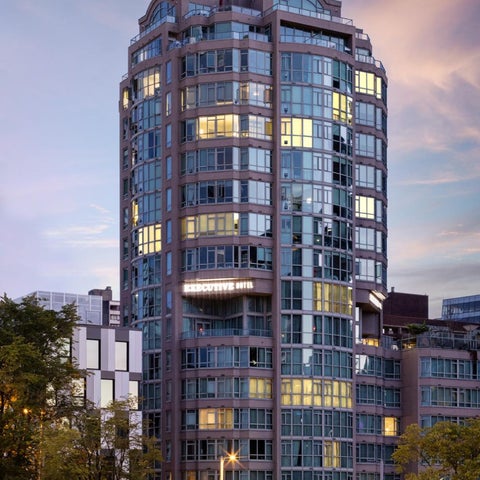 A modern high-rise building with a glass facade and illuminated windows stands against a colorful dusk sky.