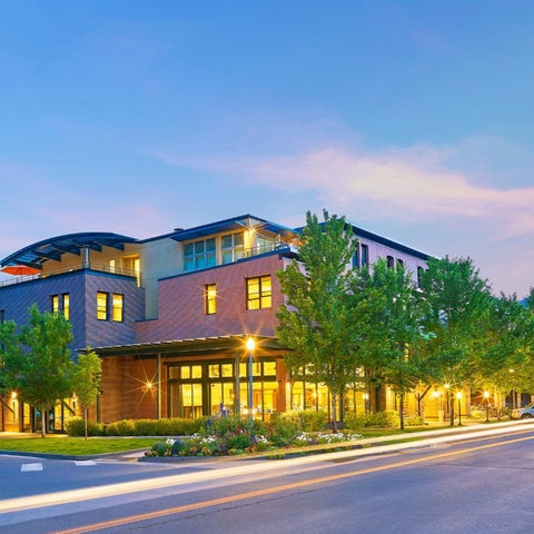A multi-story building with a modern design features a mix of brick, glass, and wood, illuminated at dusk with landscaping and streetlights.
