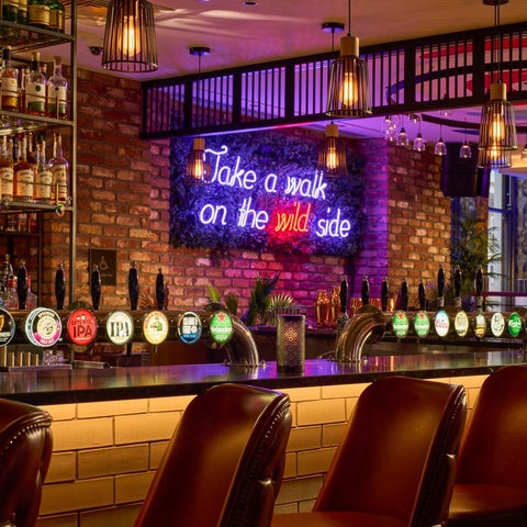 A modern bar features a long counter, dark leather stools, neon signage, and a variety of drinks and beers on tap.