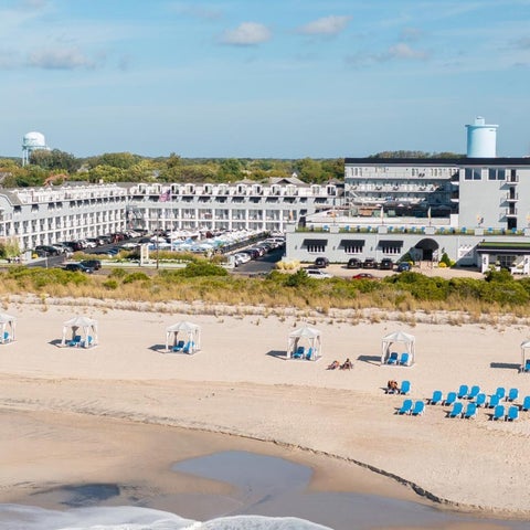 A beachfront hotel overlooks a sandy beach with lounge chairs and umbrellas under a partly cloudy sky.