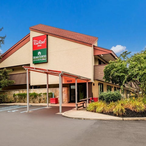 A Red Roof Inn building with a red roof and signage is shown with landscaping and a parking area in front.