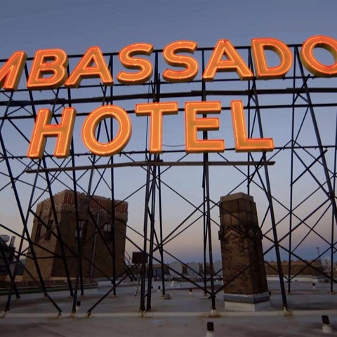 A large, illuminated 'Ambassador Hotel' sign is mounted on a metal framework against a dusky sky and surrounding buildings.