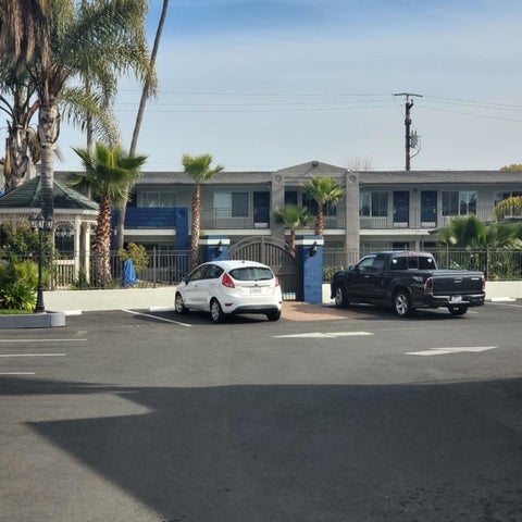 A three-story blue hotel with palm trees and a gated entrance is seen from a street with parked cars.