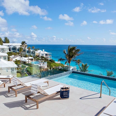 A luxury resort features a pool overlooking a turquoise ocean with lounge chairs and palm trees on a sunlit terrace.