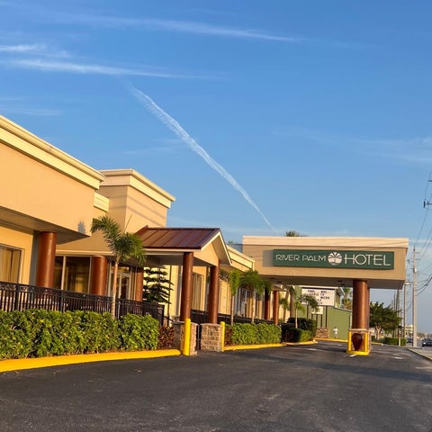 The River Palm Hotel's entrance features a tan building with a dark green sign and a driveway lined with palm trees and shrubbery.
