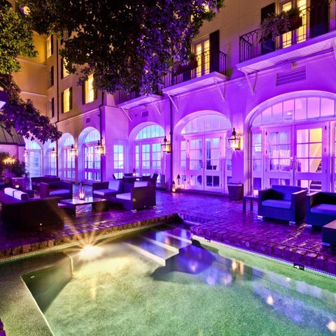 A courtyard features a pool reflecting purple and blue lighting, surrounded by lounge furniture and a building with arched windows.