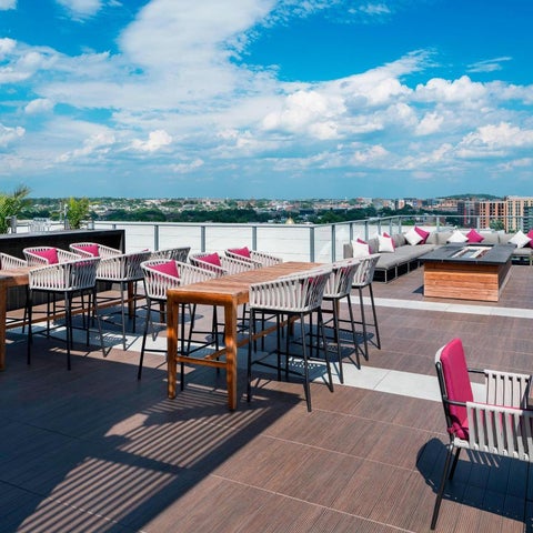 A rooftop terrace features multiple seating areas with lounge chairs, tables, and a fire pit against a cityscape backdrop.