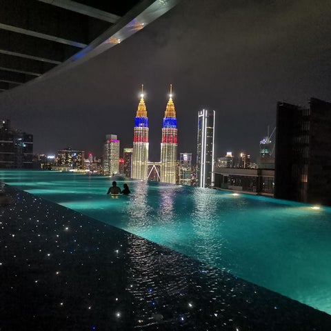 A nighttime view of a rooftop pool with the Petronas Towers illuminated in the background, featuring two people swimming.