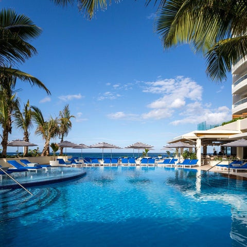 A vibrant blue pool with lounge chairs and umbrellas overlooks a turquoise ocean and a modern hotel building.