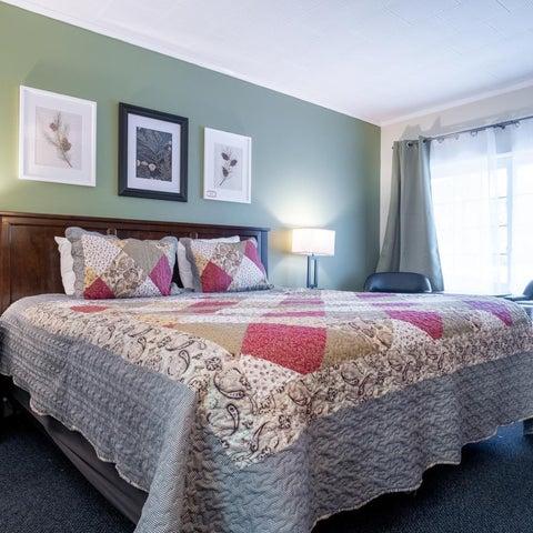 A hotel room features a king-sized bed with a patterned quilt, bedside tables with lamps, and a window with curtains.