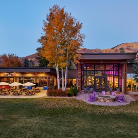 A modern building with a large patio, fire pit, lounge chairs, and lush green lawn surrounded by autumn trees.