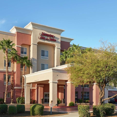 A Hampton Inn & Suites building with a covered entrance, landscaping, and palm trees in a sunny outdoor setting.