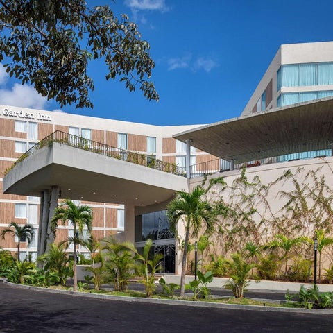 A modern hotel building with a curved overhang, lush landscaping, and palm trees against a partly cloudy sky.