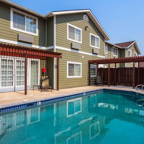 A rectangular swimming pool sits in front of a multi-story building with a covered patio and lounge chairs.