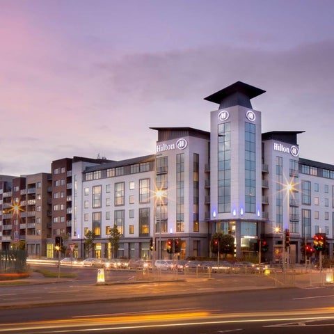 A modern hotel building with a distinctive tower facade is illuminated against a cloudy dusk sky.