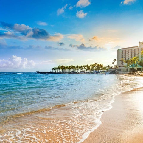 A wide view of a sandy beach with turquoise water, palm trees, and a hotel building under a partly cloudy sky.