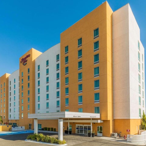 A multi-story Hampton Inn hotel building with a yellow and white facade stands against a clear blue sky.