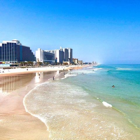 A wide view of a sandy beach with turquoise water and several large hotel buildings lining the coast under a clear blue sky.