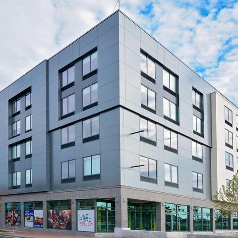 A modern, gray apartment building with large windows and a ground-level retail space stands against a partly cloudy sky.