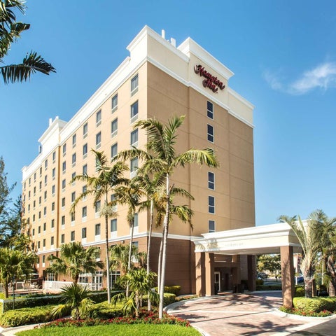 A multi-story Hampton Inn hotel building with a beige and white facade, surrounded by palm trees and landscaping.