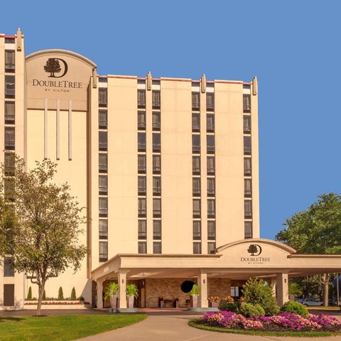 A DoubleTree hotel building with a light beige facade, a covered entrance, and landscaping with trees and flowers.