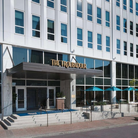 The Troubadour hotel features a modern facade with a covered entrance, patio seating, and blue umbrellas.