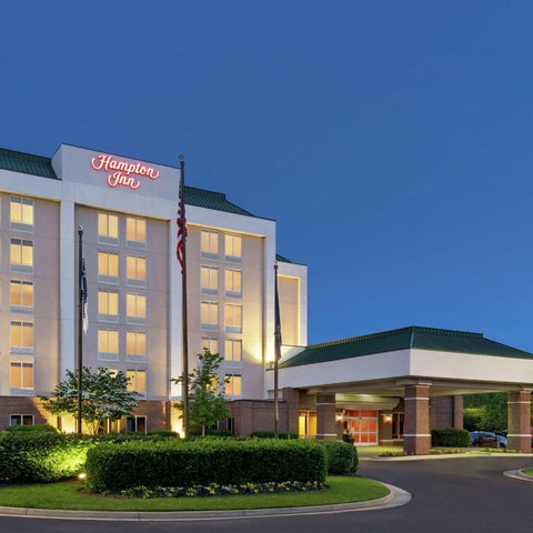A Hampton Inn hotel building is shown at dusk with a covered entrance, landscaping, and American flags.
