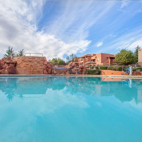 A turquoise pool with a red-orange building and landscaping reflected in the water under a partly cloudy sky.
