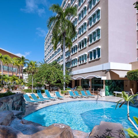 A turquoise pool with lounge chairs and palm trees sits in front of a multi-story hotel building with a light-colored facade.