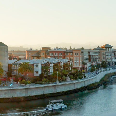 A waterfront view of a multi-story building complex with a tower, palm trees, and a river with a boat in the foreground.