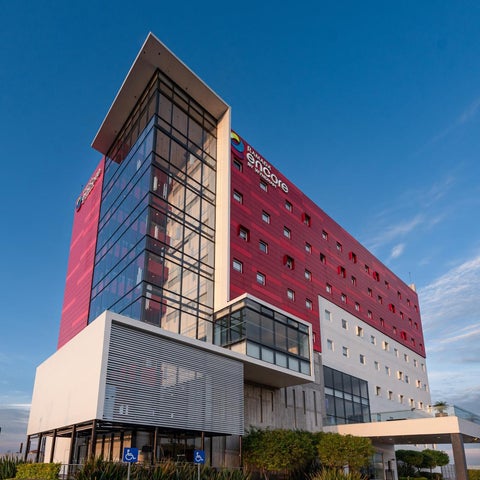 A modern, multi-story hotel building with a red and white facade and large glass windows is shown against a blue sky.