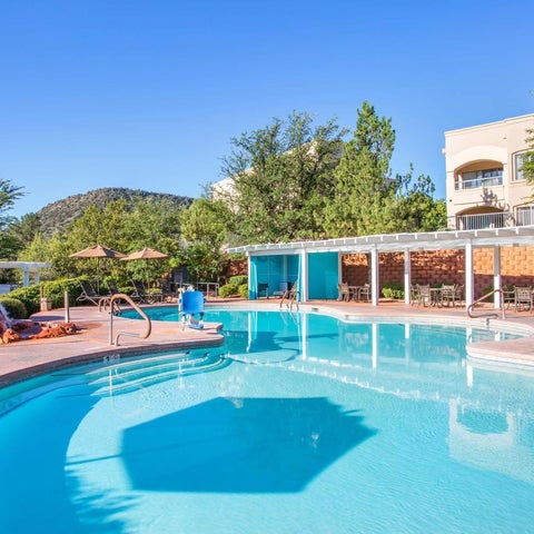 A bright blue pool with a curved edge is surrounded by patio furniture and lush greenery under a clear blue sky.