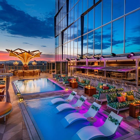 A rooftop pool area features lounge chairs, a sleek pool, and a decorative chandelier against a vibrant sunset sky.