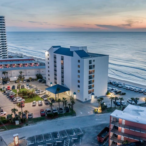 An aerial view shows a beachfront hotel complex with multiple buildings, parking, and a road along the coastline at sunset.