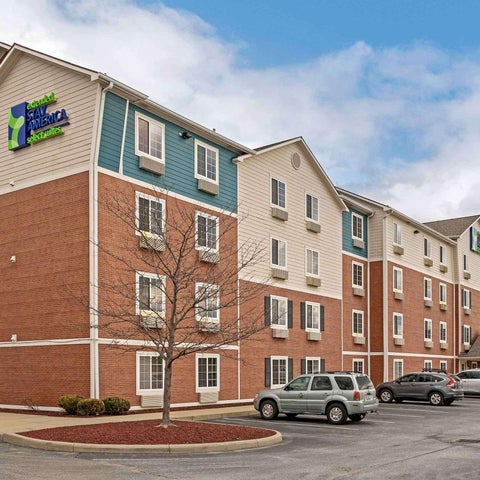 A three-story Staybridge Suites hotel building with a brick and stucco facade and a parking lot with several cars.