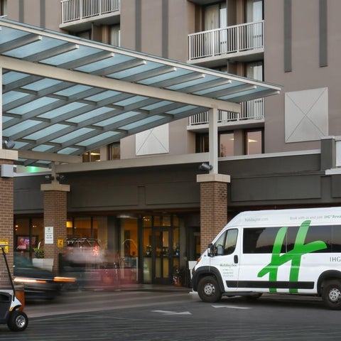 A hotel entrance features a covered drop-off area with a shuttle van and several cars parked nearby.