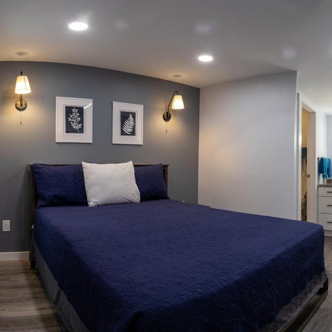 A bedroom features a dark blue bed with decorative pillows, wall-mounted lamps, and framed artwork against a gray accent wall.
