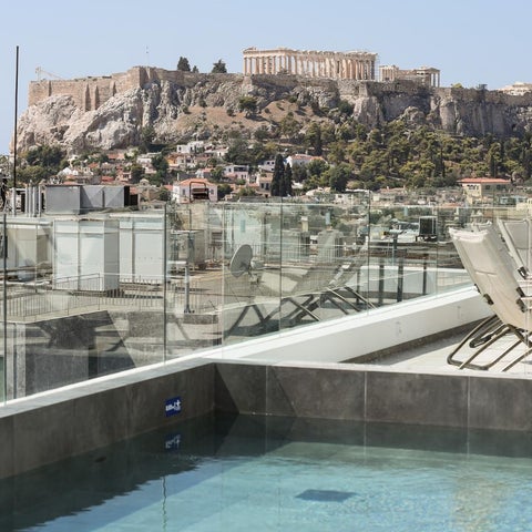 A modern rooftop pool with a glass railing overlooks the Acropolis and surrounding cityscape under a clear sky.