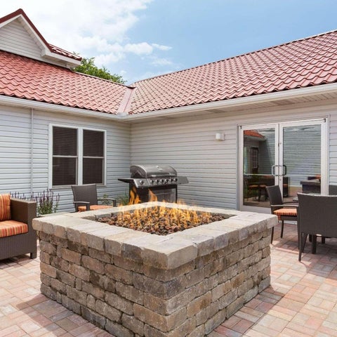 A patio area features a stone fire pit, outdoor dining set, lounge chairs, and a grill against a building with a red tile roof.