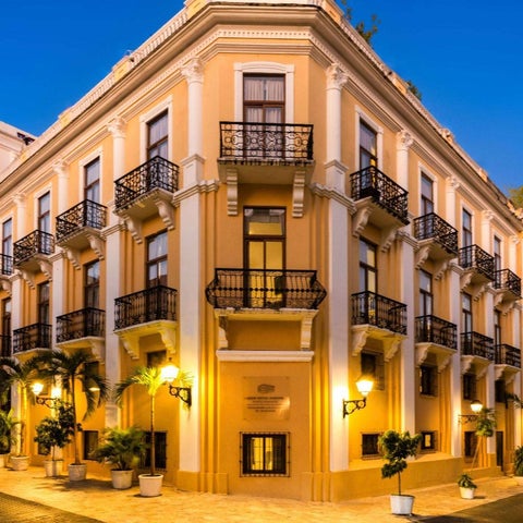 A multi-story building with ornate balconies and a cream-colored facade stands on a street lined with palm trees and lampposts.