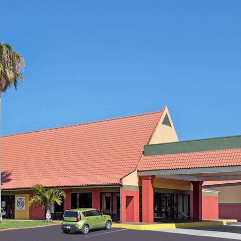A two-story hotel with a red tile roof and a covered entrance features a palm tree and a green car in the foreground.