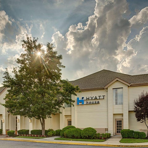 A Hyatt House hotel building facade is shown with a tree and landscaping in front of it under a cloudy sky.